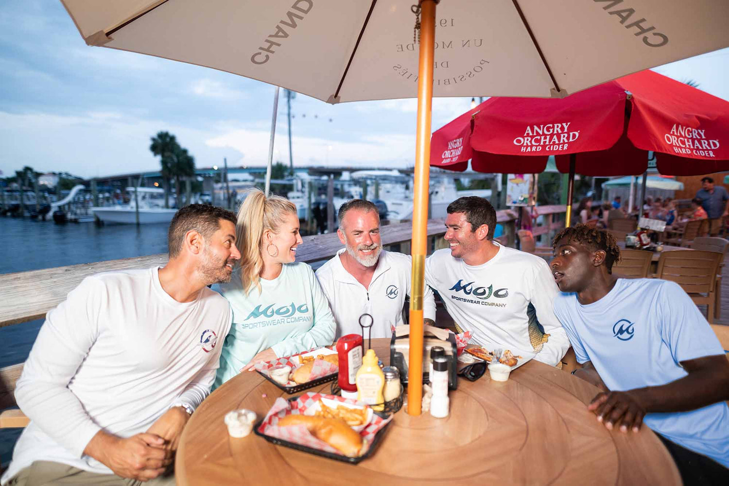 Group of 4 men and 1 woman wearing MSC performance gear and sitting around an outdoor table with food. In the background is a harbor with boats docked in the water